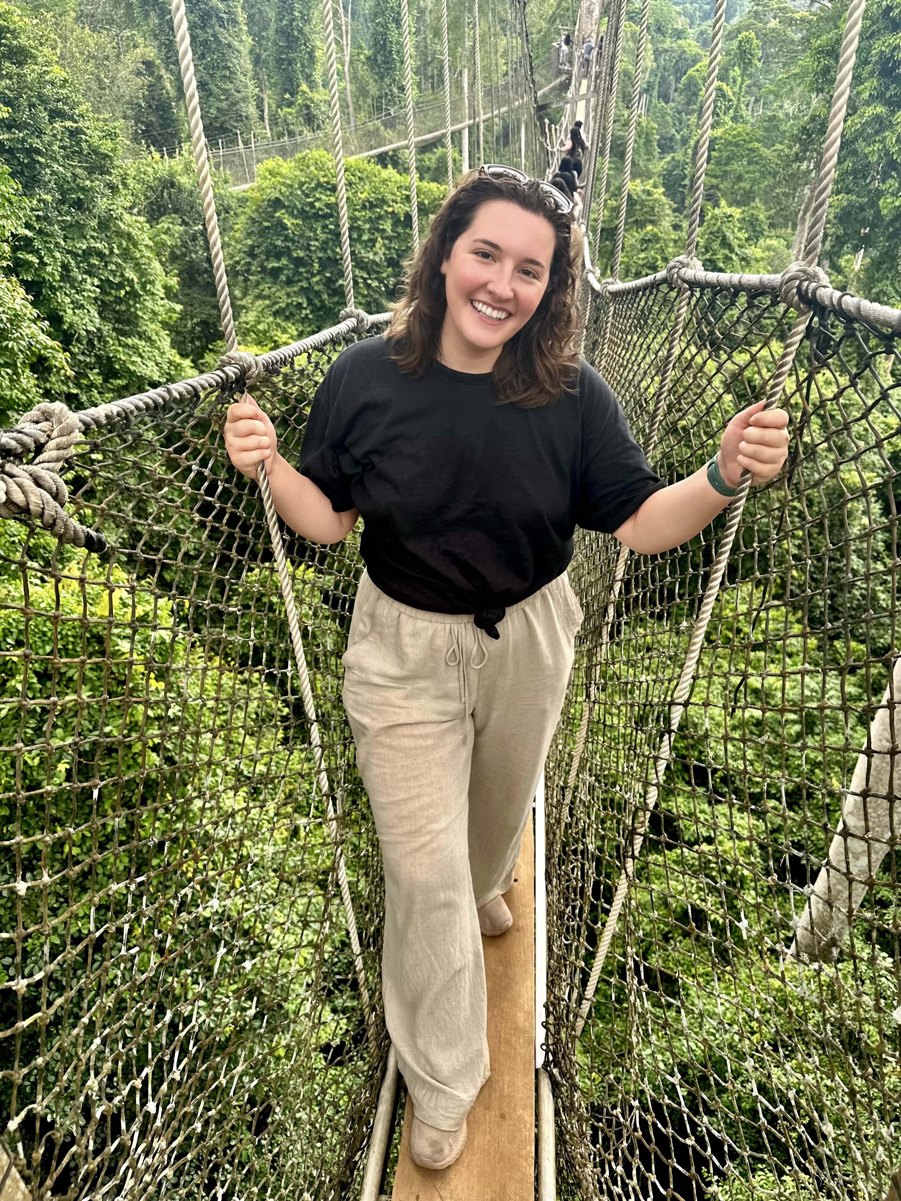 a woman wearing a black tshirt and khaki pants standing on a bridge made of ropes high above the trees in Ghana