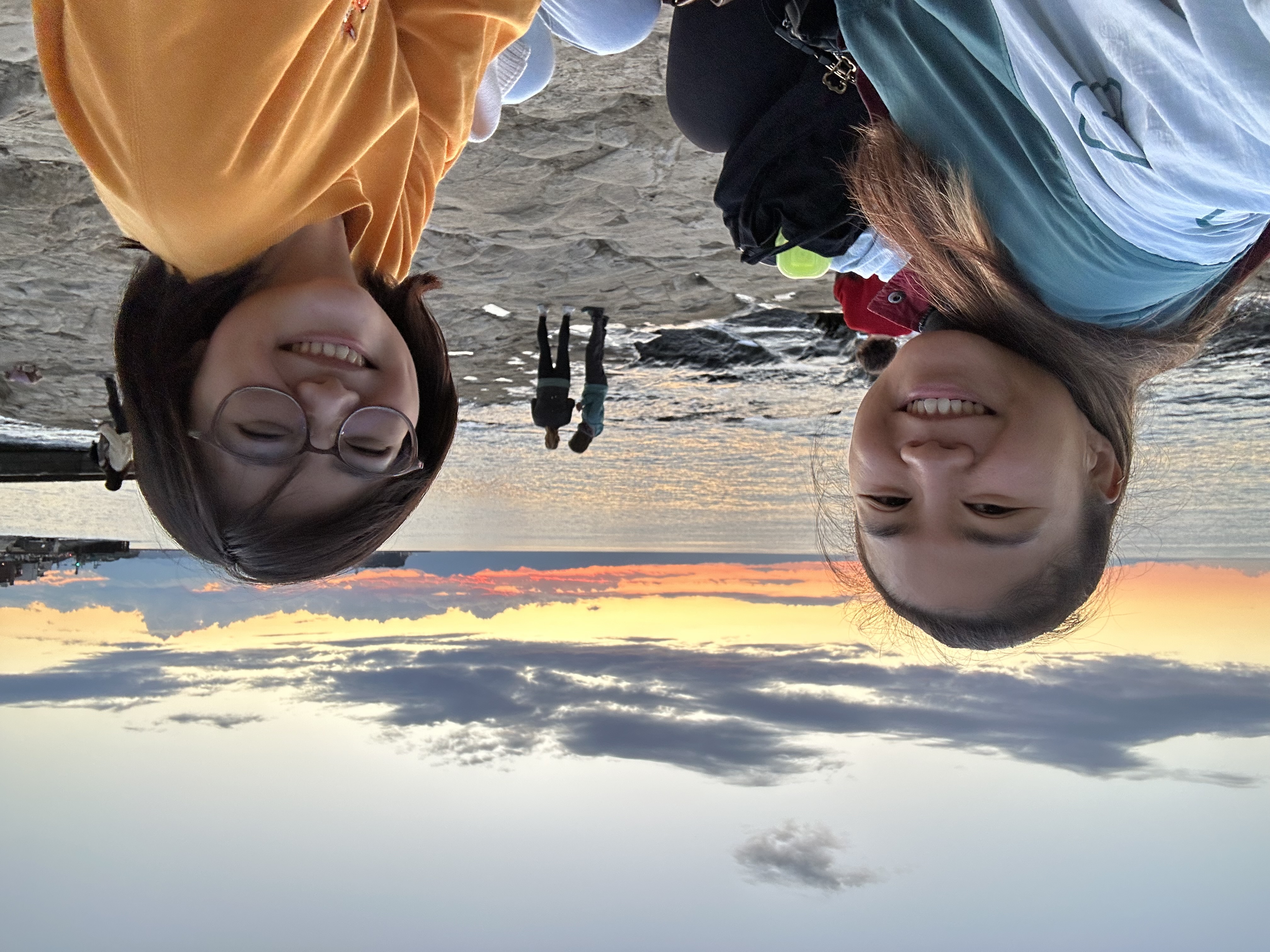 student studying abroad in Japan wearing a yellow crewneck and glasses sitting on a beach with a friend with a sunset in the background
