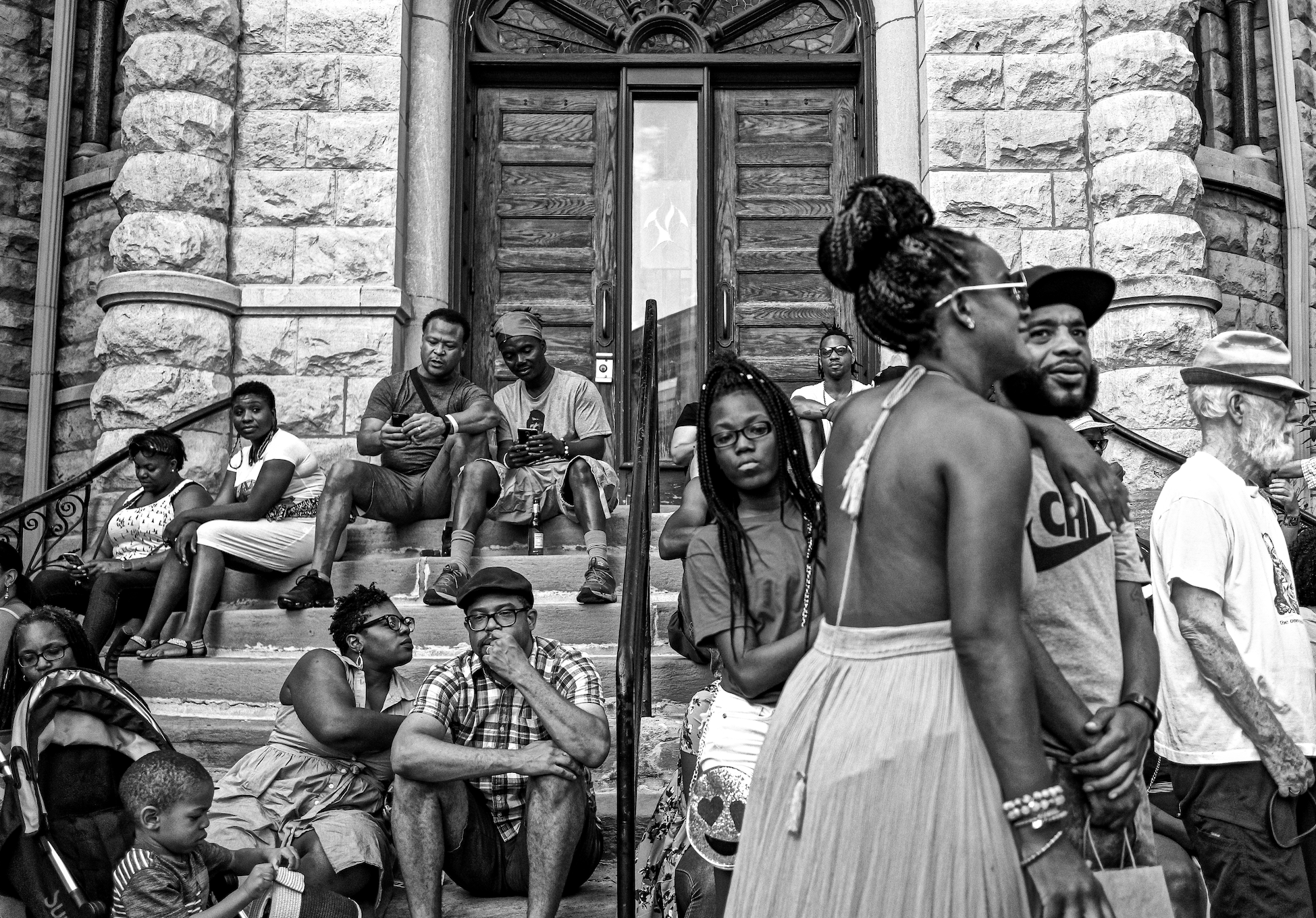 group of 16 people; some sit on steps in front of building while others walk by